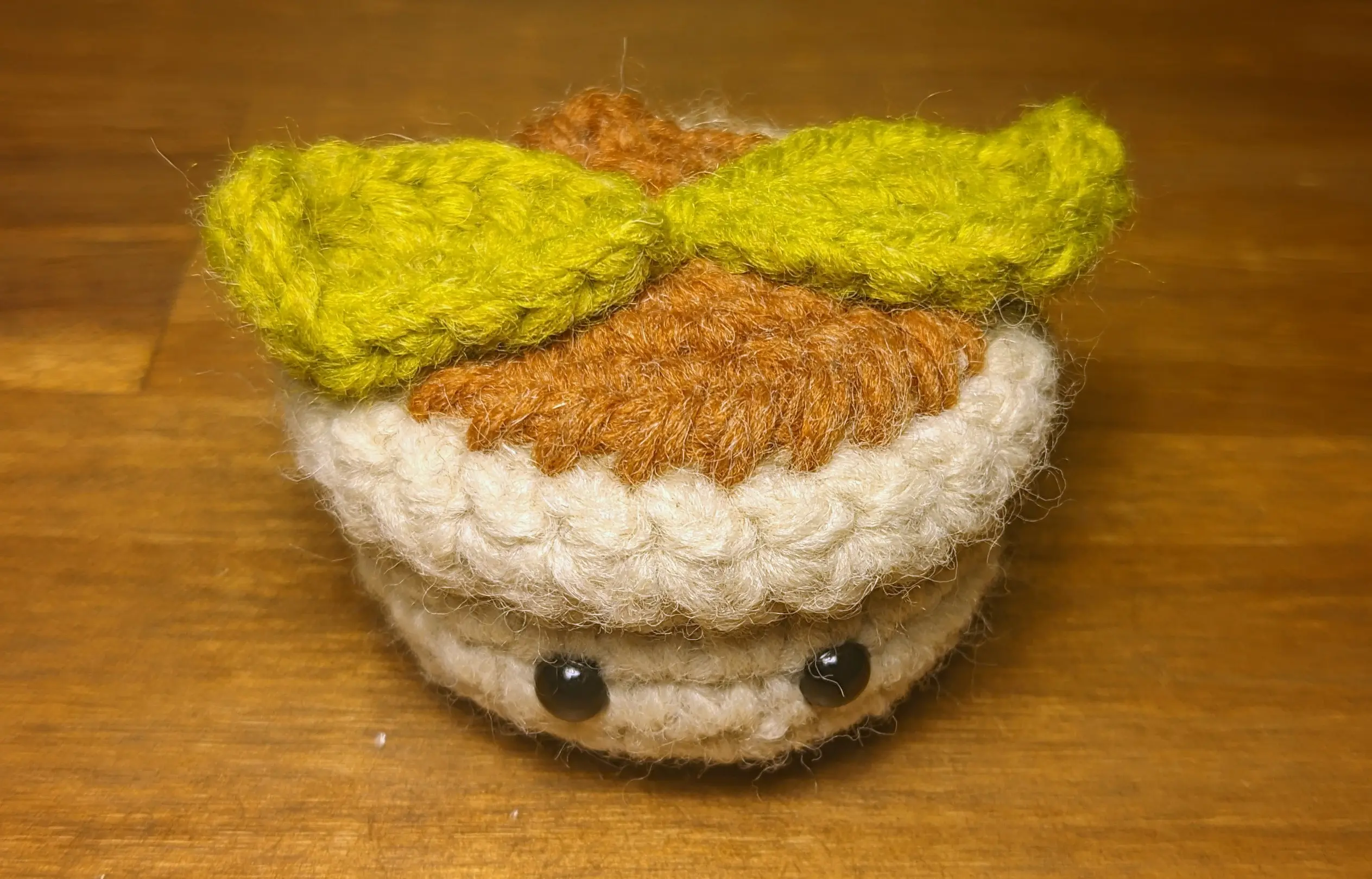 Small crochet flowerpot looking at the camera.
Two oval leaves have been fixed to the top, spanning from the left side of the pot to the right. The outward-pointing tips of the leaves curl upwards slightly, giving the leaves more dimension.