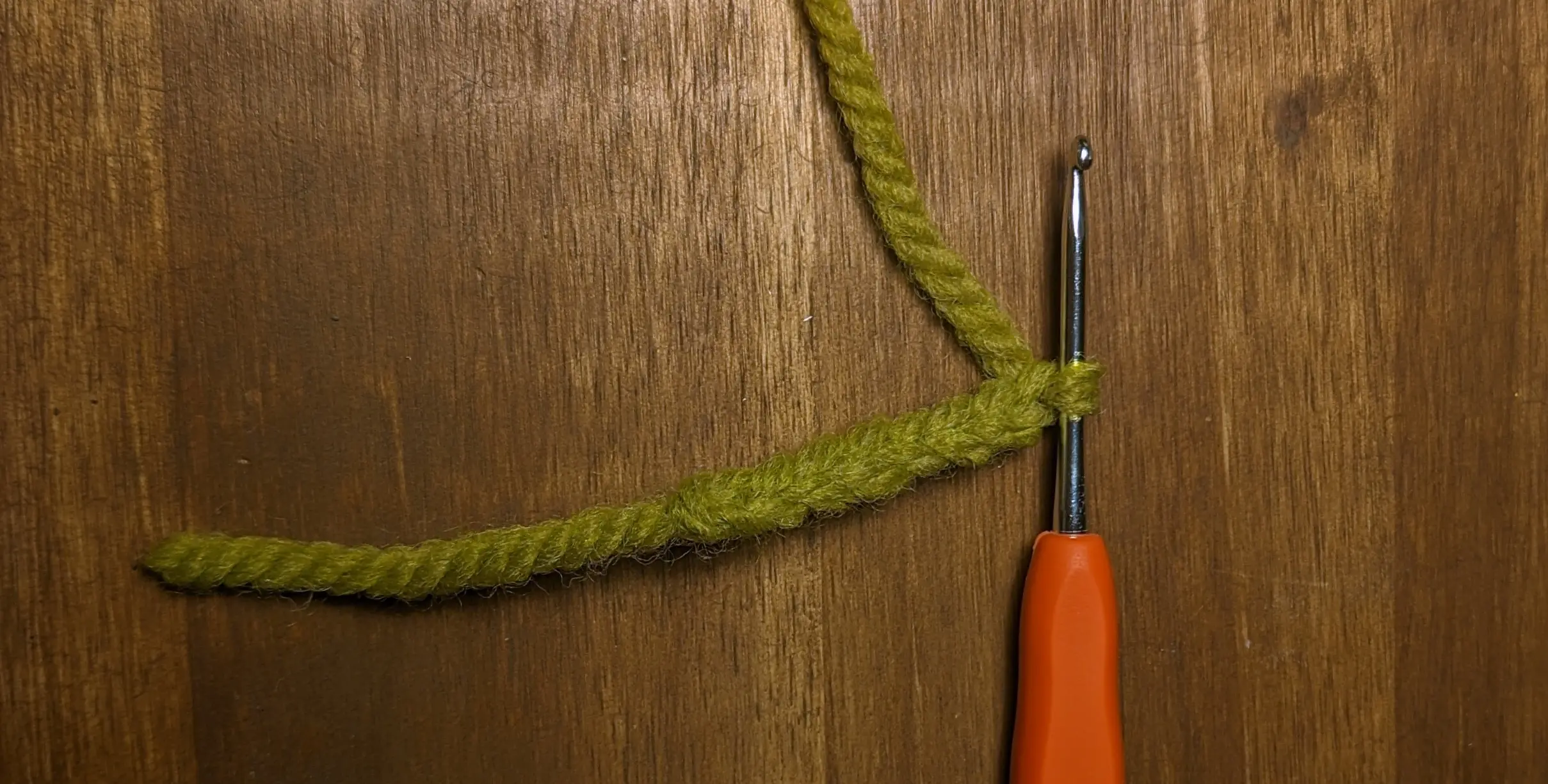 Short crochet chain made from green yarn on an orange hook. The active yarn is going off-screen, while the yarn not at the beginning of the chain is only about 4 cm long.