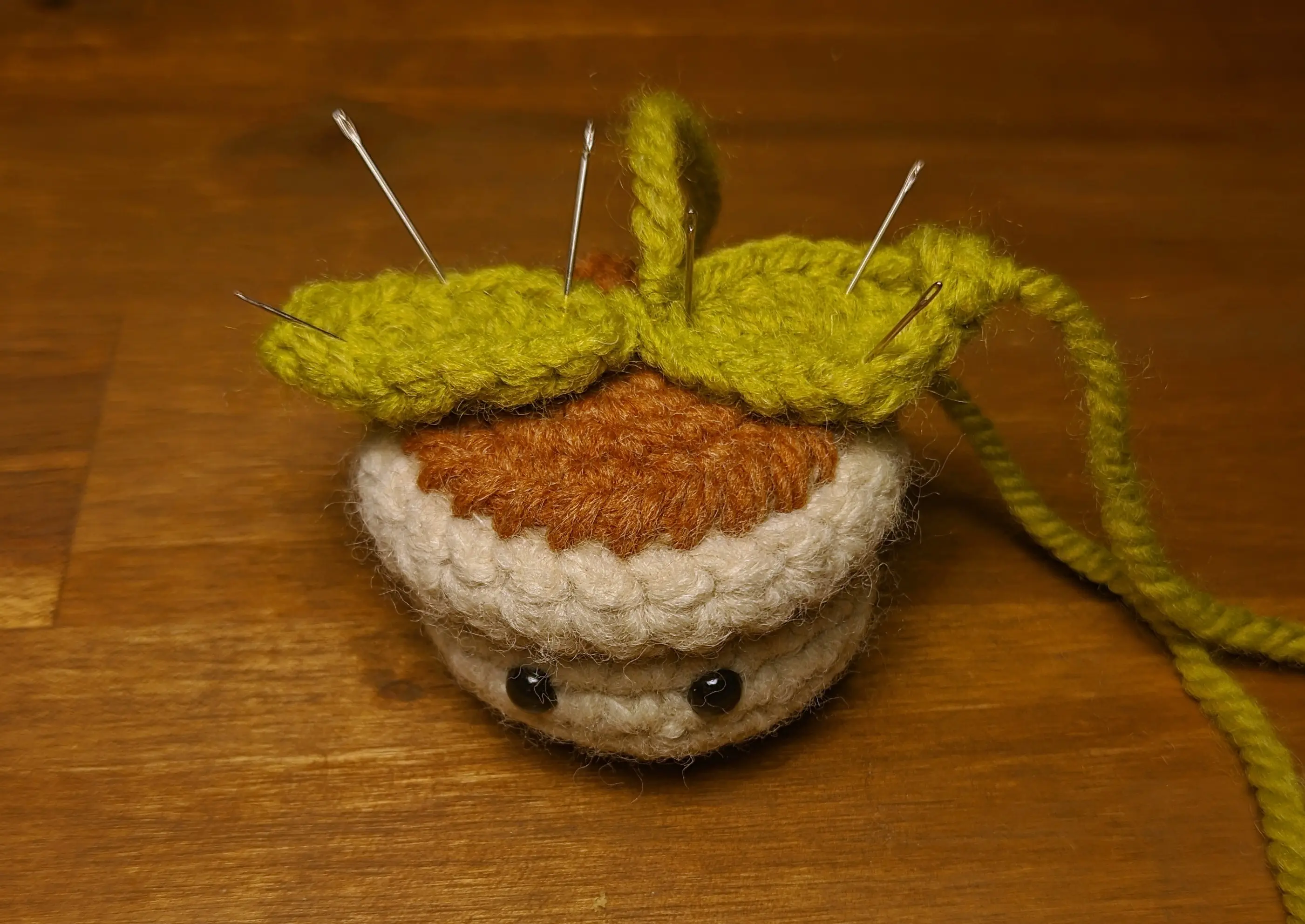 A tiny crochet flowerpot with black eyes placed on a desk.
Two green leaves have been pinned on the top using sewing needles. The excess yarn is just running off the sides.
One leaf starts in the middle and slightly overhangs on the left of the pot. The other leaf starts in the middle and slightly overhangs on the right of the pot.