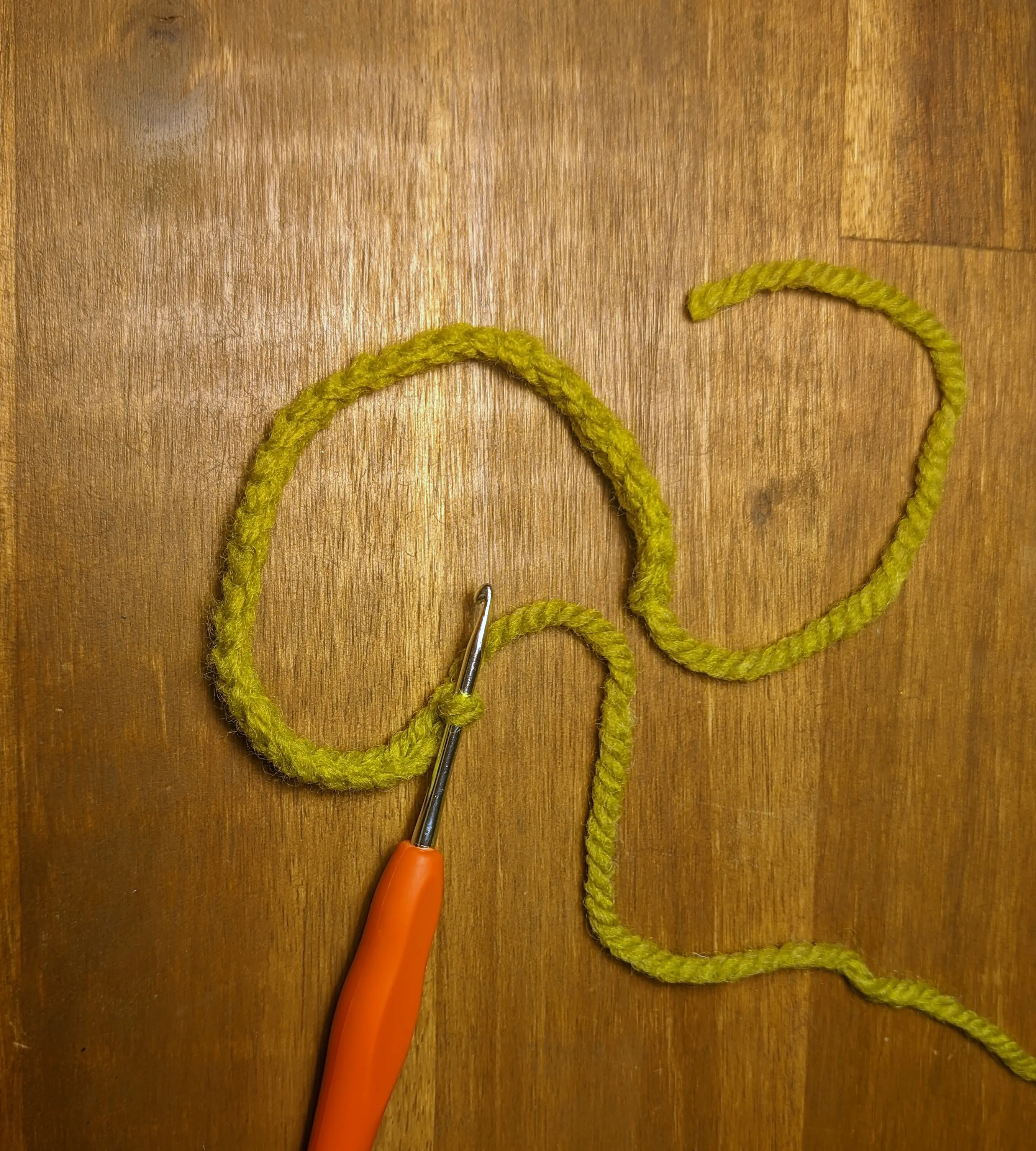 A long curled-up crochet chain on a wooden desk. The crochet hook is stuck in the last loop of the chain.