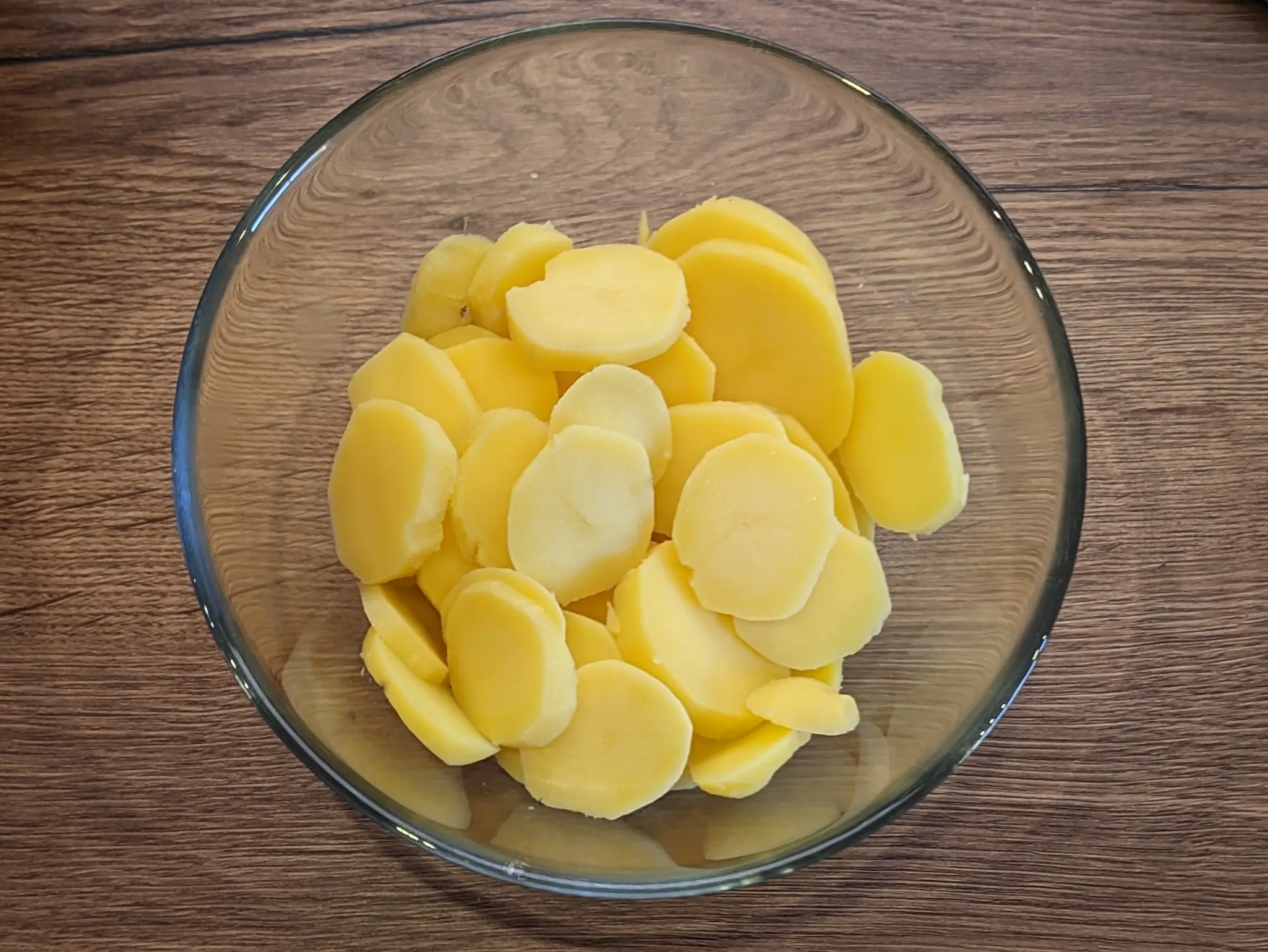 Top-down shot of potato slices in a glass bowl on a wooden work surface.