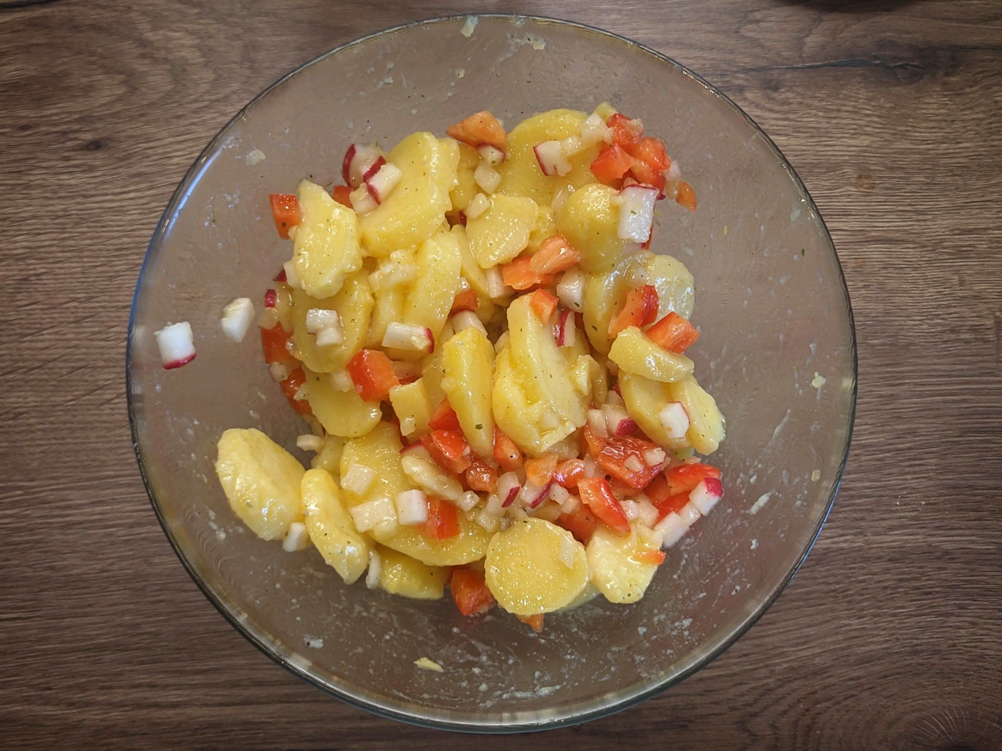 Top-down shot of the same glass bowl, but now the potatoes have been mixed with small cubes of red bell pepper and radishes.