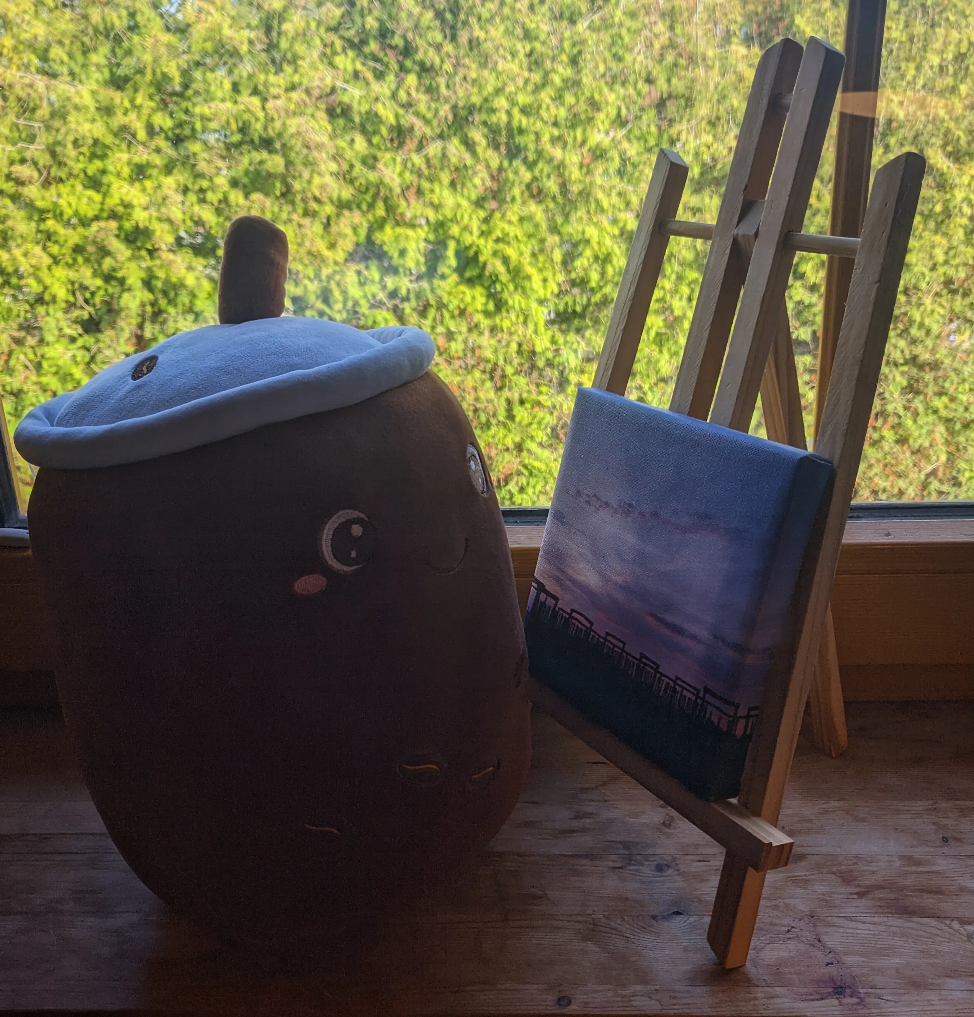 Dark brown bubble tea plushy sitting on a window sill in front of a canvas on an easel. Out the window the sun lights up green trees.